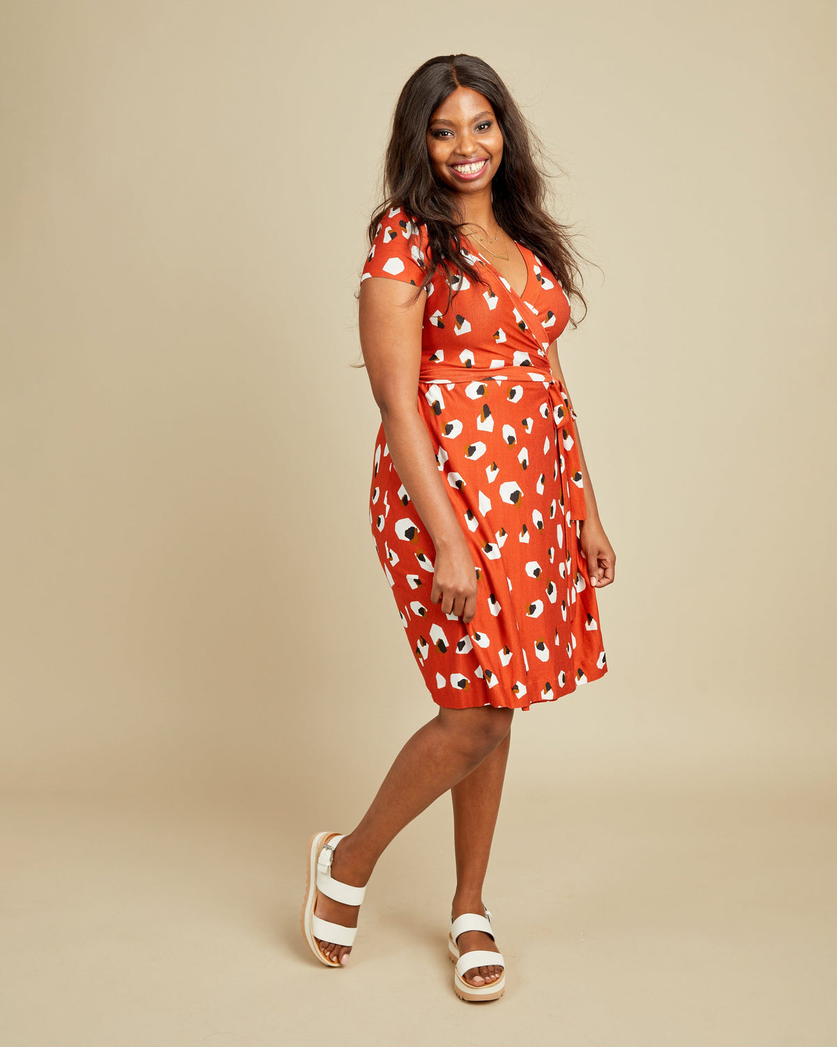 Woman wearing a red dress with white patterns on a beige background. This dress is the Appleton Wrap Dress sewing pattern from Cashmerette.