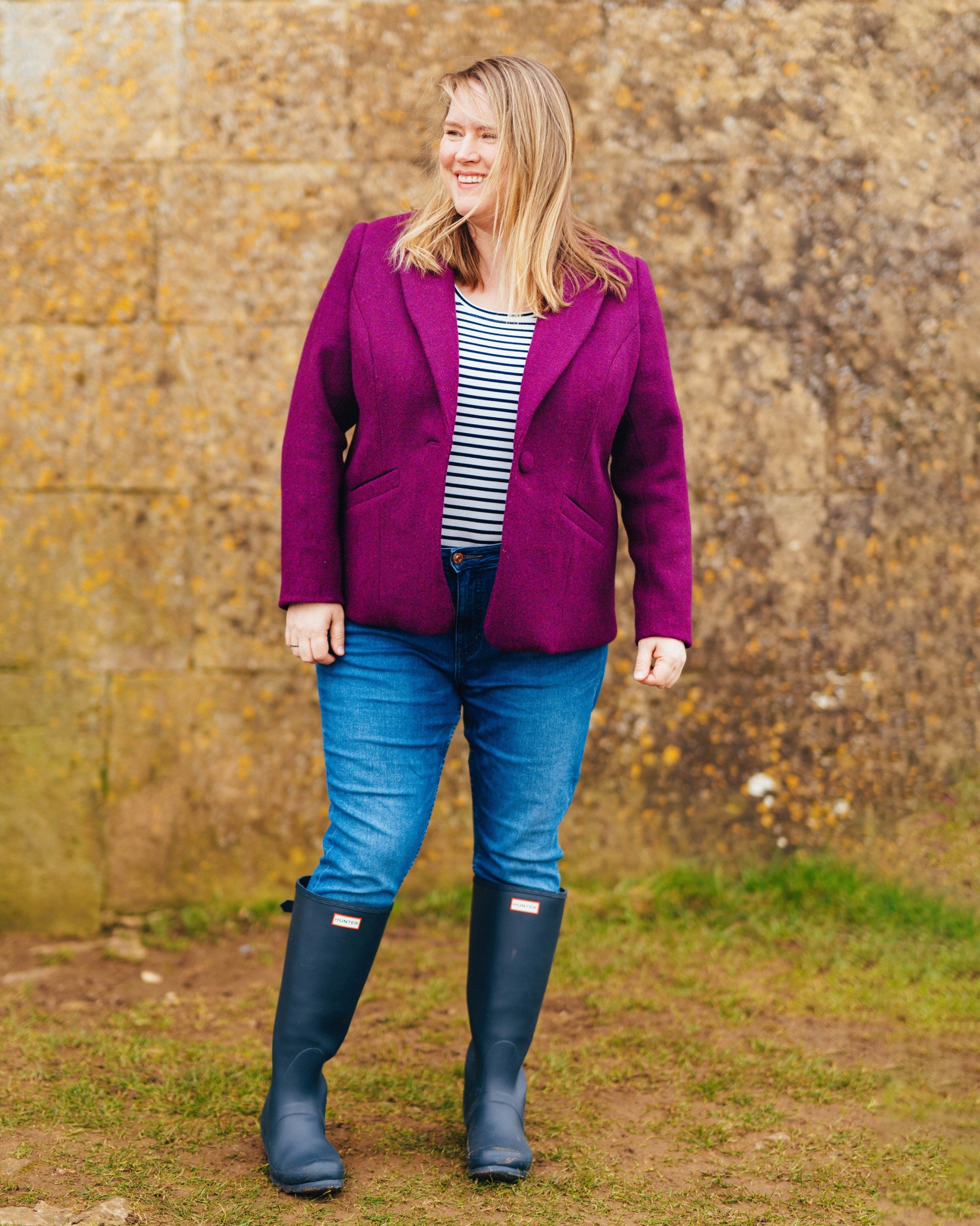 Woman wearing a purple jacket, striped shirt, blue jeans, and navy boots standing in front of a stone wall. This blazer is the Auburn Blazer sewing pattern from Cashmerette.