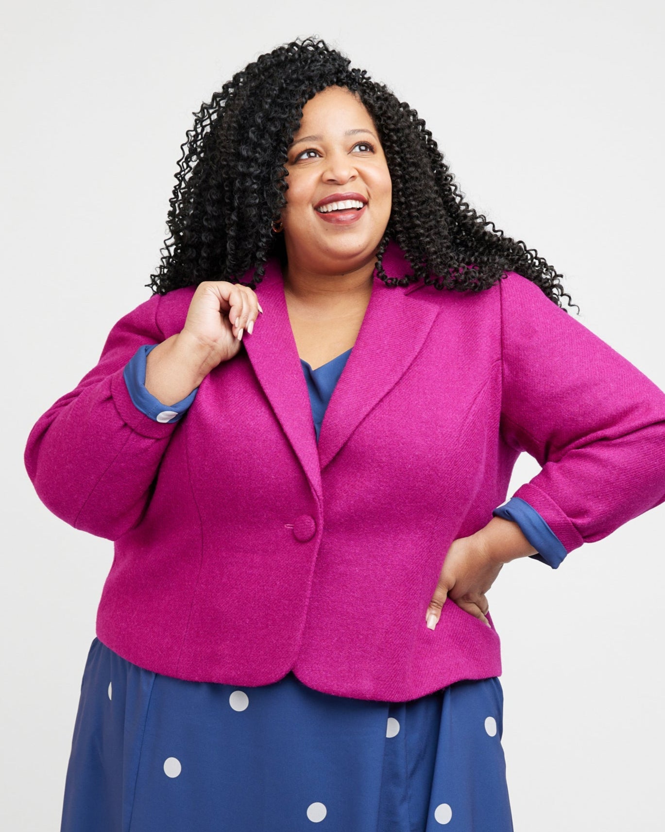Woman wearing a bright pink blazer over a blue dress with white polka dots on a white background. This blazer is the Auburn Blazer sewing pattern from Cashmerette.
