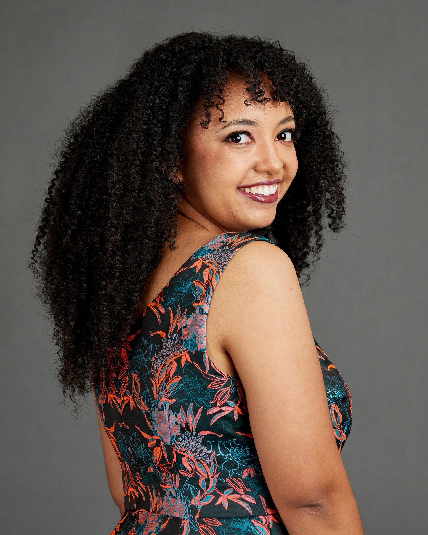 Woman with curly hair wearing a floral dress against a gray background. This dress is the Upton Dress & Skirt sewing pattern from Cashmerette.